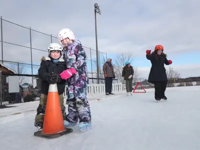 three children skating on outdoor ice pad, two of which holding pylon