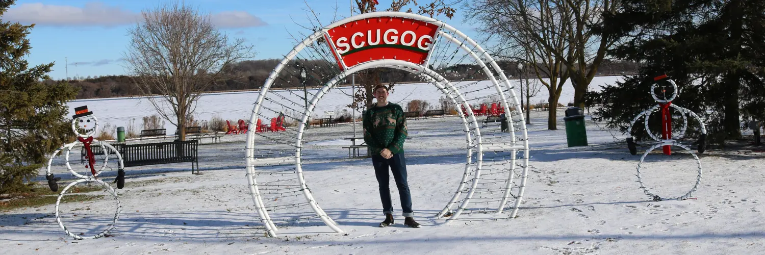 Man in green Christmas sweater standing under metal 'Scugog' archway in snowy Palmer Park. Metal snowman sculptures are stationed on either side of archway.