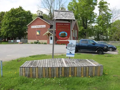 Front of Scugog Shores Museum Village with schoolhouse in background and forefront sign naming the Museum