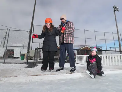 man helping child onto outdoor ice pad with another child sitting on side of rink