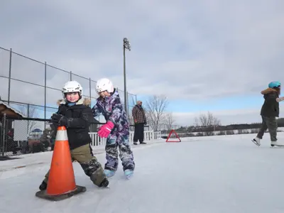 two children skating on outdoor ice pad holding pylon